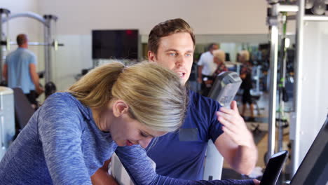 Mujer-Haciendo-Ejercicio-En-La-Máquina-De-Ciclismo-Siendo-Alentada-Por-Un-Entrenador-Personal-En-El-Gimnasio.