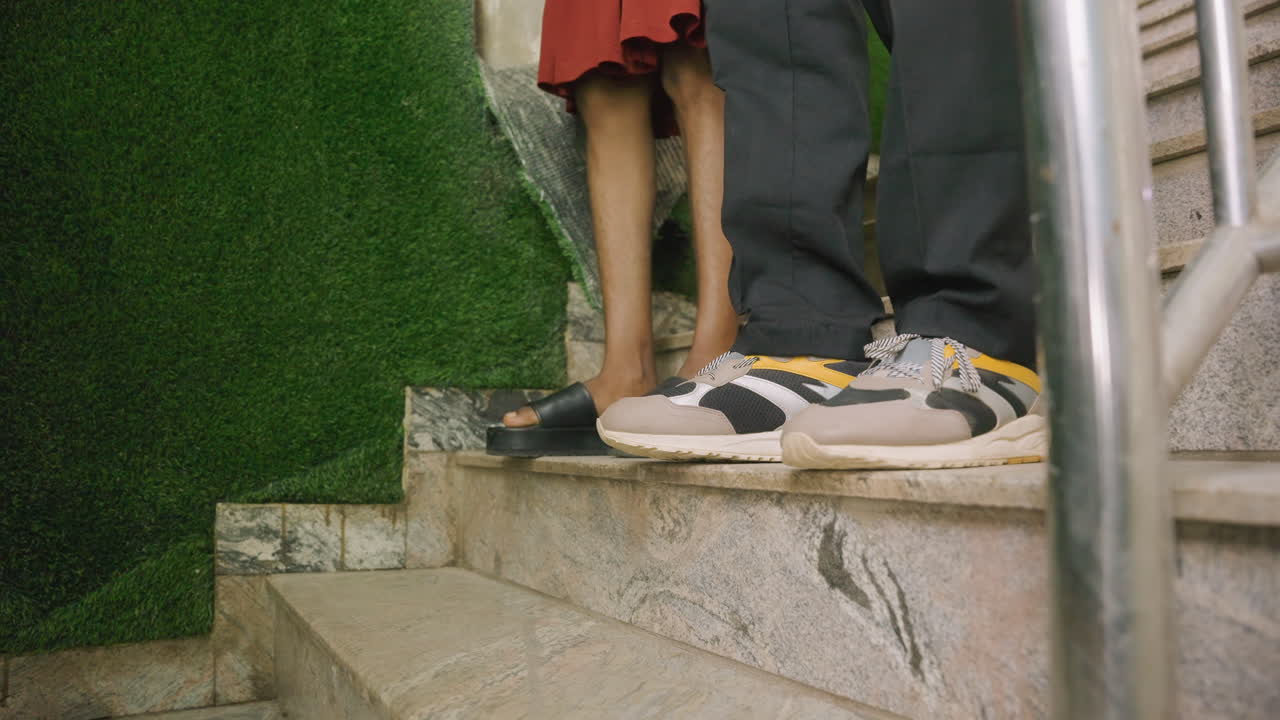 Close-up kaki pasangan di tangga pada kencan intim, menunjukkan pria bersneaker dan wanita bersandal, tangga marmer di samping dinding hijau, sentuhan lembut antar pasangan, butiran film dan suasana malam yang hangat.