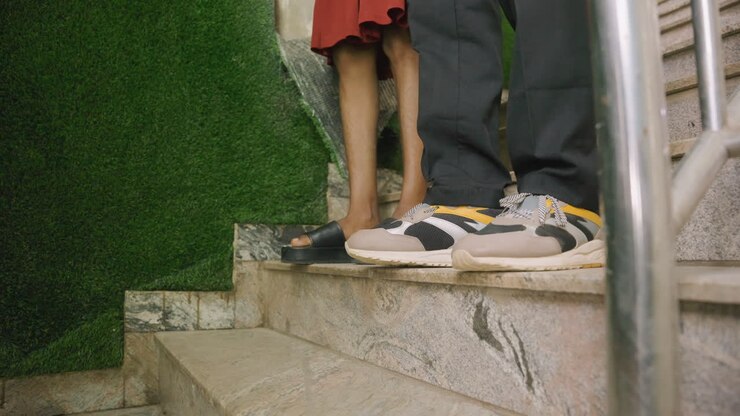 Primo piano delle gambe di una coppia su una scala durante un appuntamento intimo, mostrando l'uomo in scarpe da ginnastica e la donna in sandali, gradini di marmo accanto a una parete verde, tocco delicato tra i partner, grana della pellicola e atmosfera serale calda.