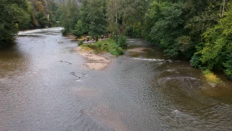 drone rises over a group riding on horses through a river by a castle