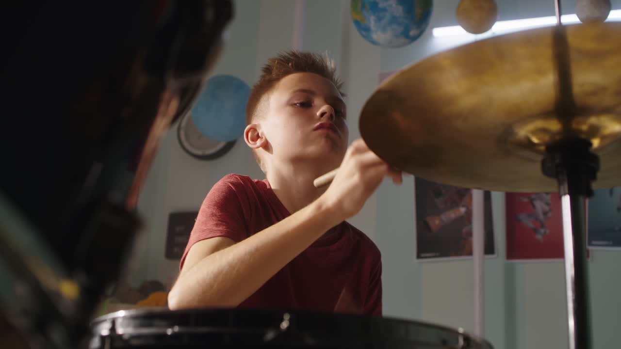 Teenager Playing Drums at Home from below serious Teen Musician in Red T Shirt Practicing to Play Drums during Music Session at Home