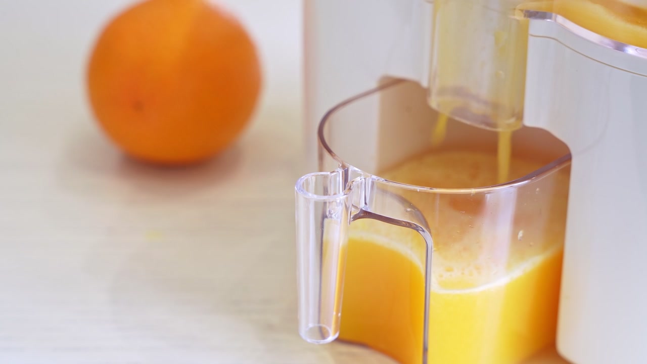 Nærbillede af frisk appelsinjuice, der hældes fra en hvid elektrisk juicer ned i en gennemsigtig beholder. En hel appelsin ligger i baggrunden af et lyst, moderne køkken om dagen.