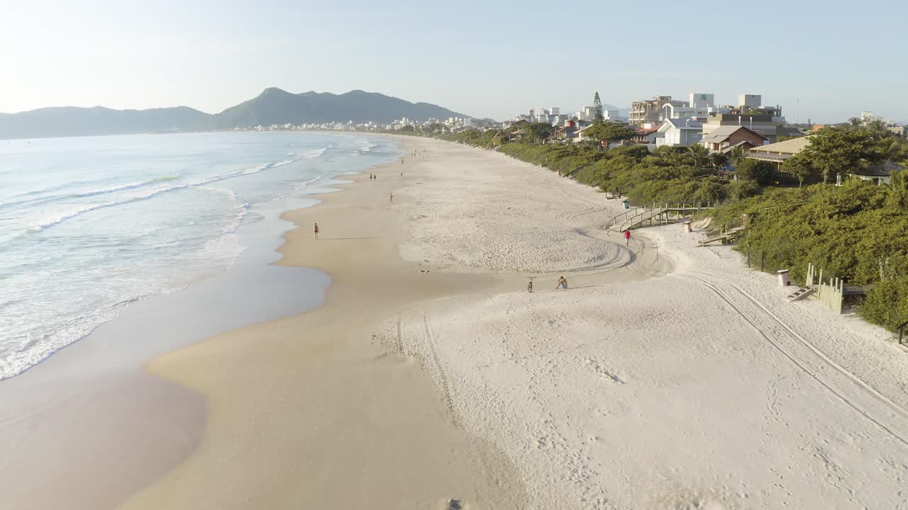 Drone voando perto de uma bela praia com ondas - Paisagem costeira com montanhas e pequenos edifícios ao fundo de uma praia paradisíaca tropical com areia branca e águas azuis do oceano brasileiro