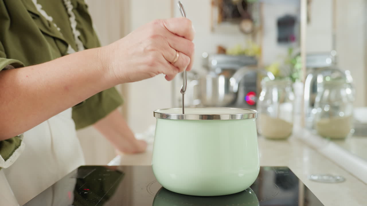 Chef wearing green top and white apron stirs food inside pastel green pot on electric stove in bright home kitchen, focusing on hand movement and utensil use with modern appliances