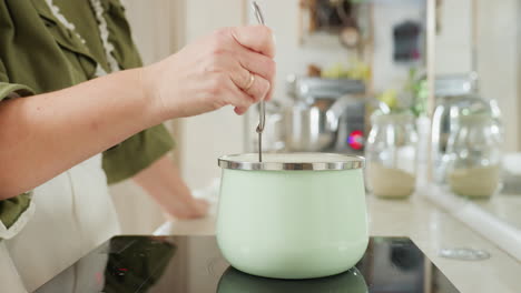 chef wearing green top and white apron stirs food inside pastel green pot on electric stove in bright home kitchen, focusing on hand movement and utensil use with modern appliances