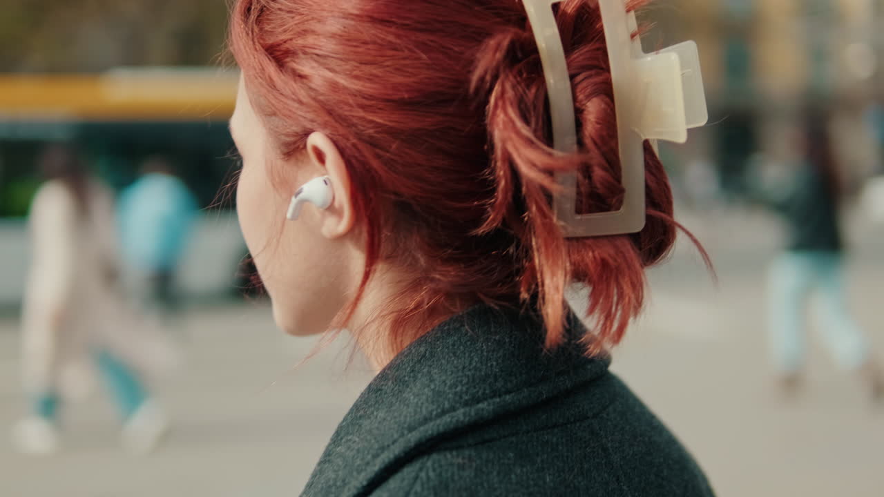 Giovane donna dai capelli rossi che ascolta la musica con gli auricolari wireless in città