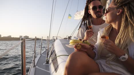 stylish couple with dreadlocks in white clothes and sunglasses sitting embracing on the edge of the yacht smiling, clinking with champangne glasses. loving couple spending time together on the yacht beyond the sea