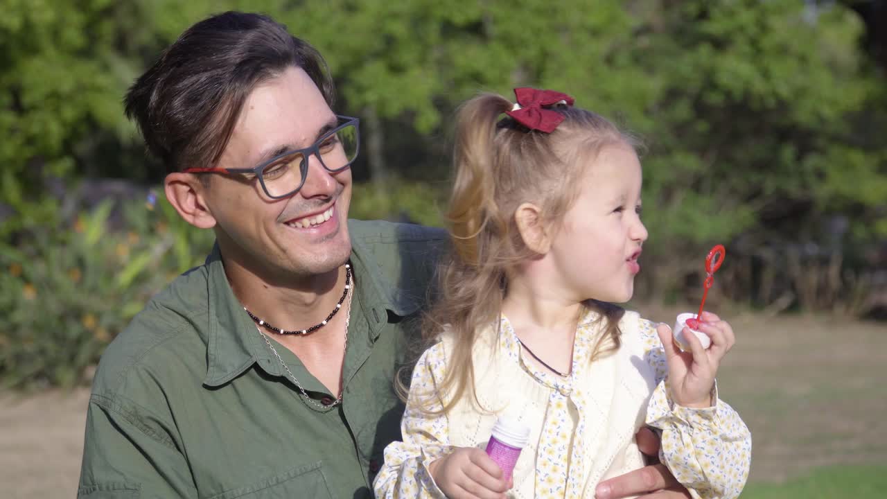 Un giovane padre sorridente tiene in braccio la sua adorabile figlioletta mentre lei gioca con le bolle nel parco cittadino.