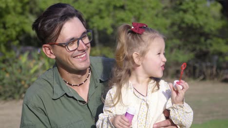 un joven padre sonriente sostiene a su linda hija mientras ella juega con las burbujas en el parque de la ciudad