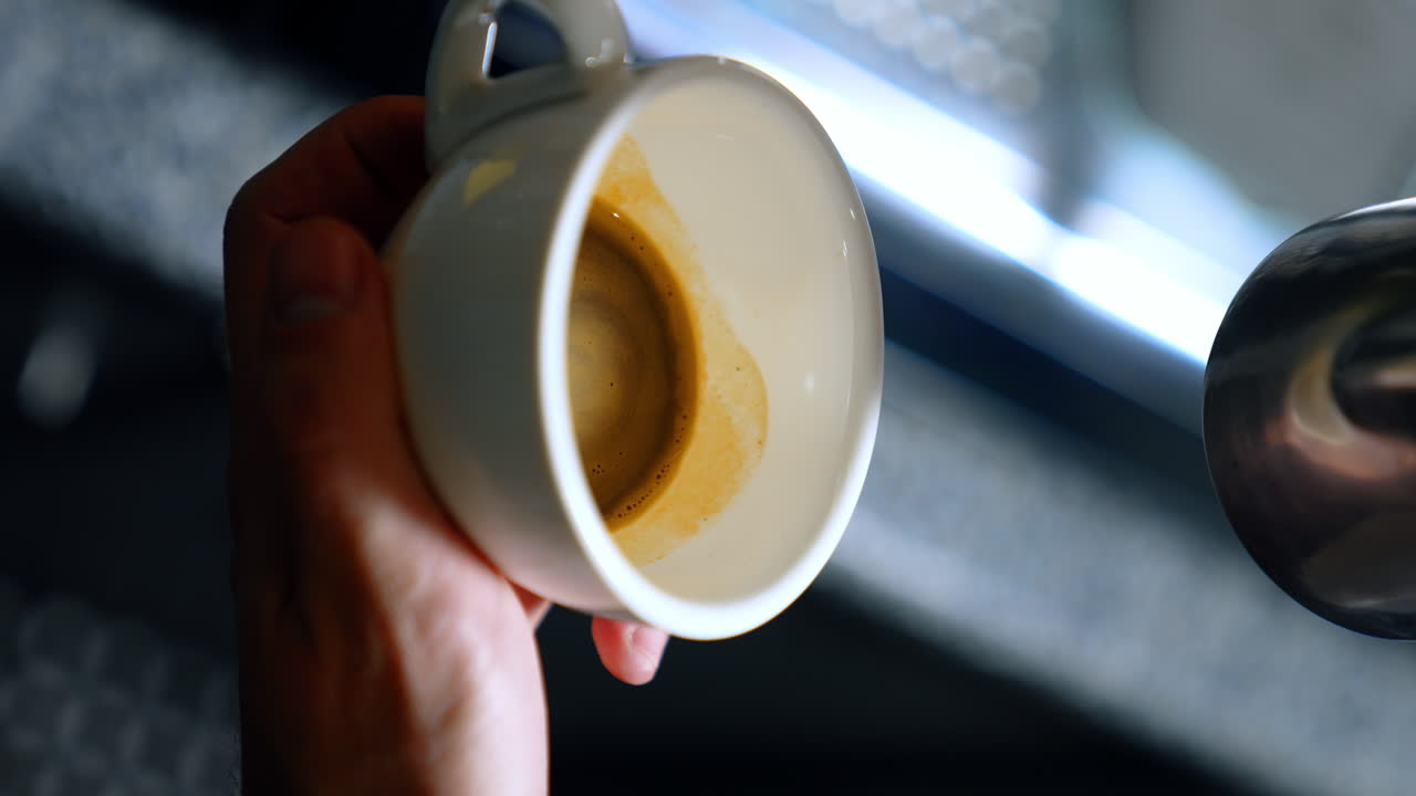 Male hands hold a white coffee cup filling it with milk. Barista preparing latte drink. Close up. Vertical screen. Blurred backdrop.