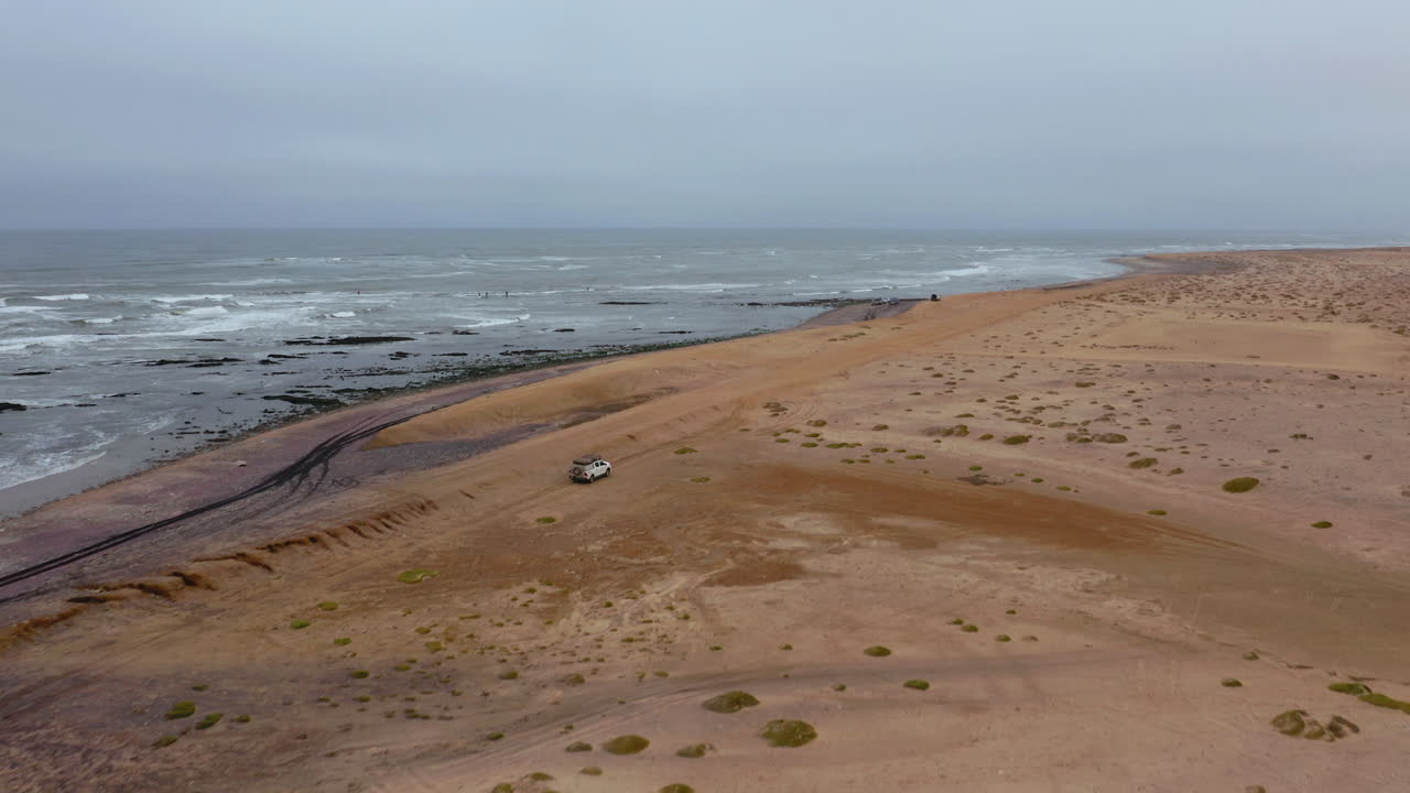 Slow drone reveal shot of a lone vehicle driving on a vast, empty beach near Swakopmund on a moody, overcast day
