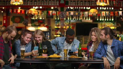 group of friends of bar students drink beer and look at the screens of smartphones smiling.