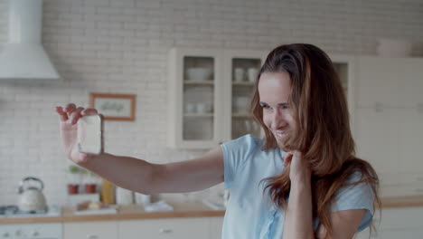 Cheerful-woman-winking-to-phone-camera-in-kitchen.-Girl-taking-selfie-photo.