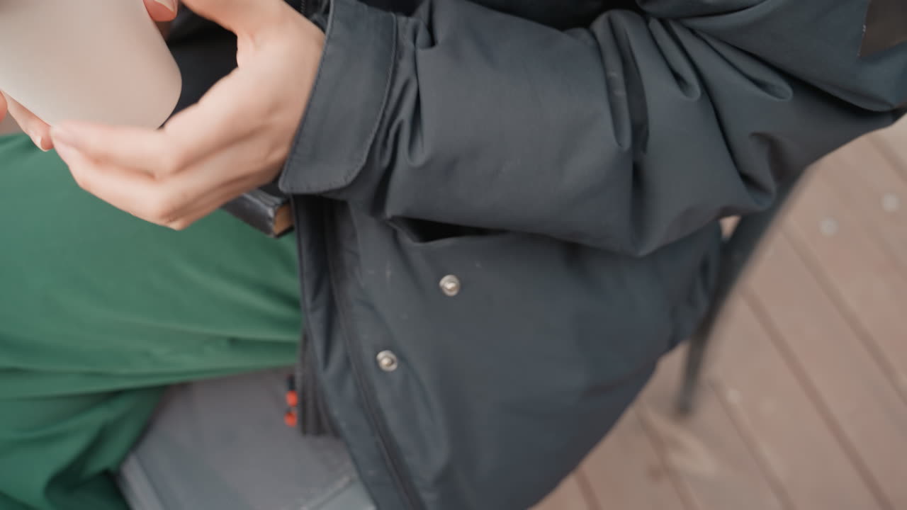 hands holding disposable coffee cup outdoors, winter jacket, green pants, wooden terrace table, warms hand in pocket, places cup on table, secures lid, freelance writer taking break, student flipping