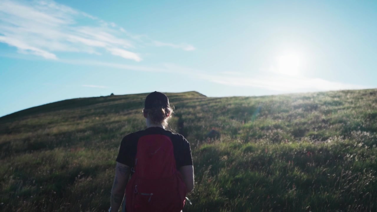 Following A Woman Wearing Cap And Backpack While Trekking On Grassy Hill With Pet Dog On A Sunny Day. medium shot