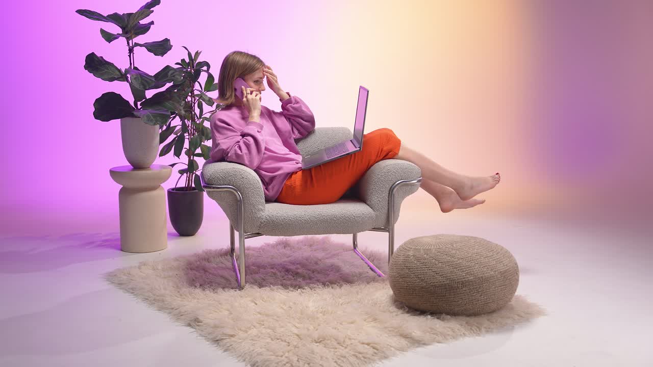 A relaxed young woman sits barefoot in a modern armchair using a laptop and talking on a smartphone. The pastel pink and orange background with decorative plants adds a soft, contemporary atmosphere