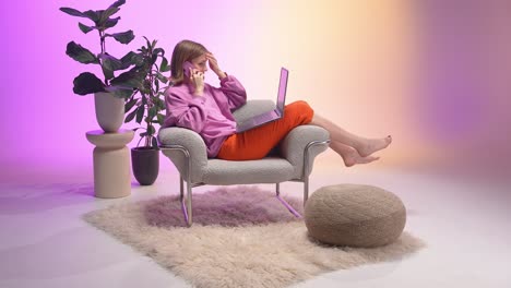 a relaxed young woman sits barefoot in a modern armchair using a laptop and talking on a smartphone. the pastel pink and orange background with decorative plants adds a soft, contemporary atmosphere