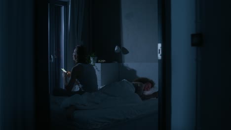 couple in bedroom at night, one is using a phone while the other is sleeping