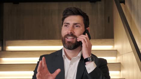 positivo hombre barbudo sonriente hablando por teléfono móvil en las escaleras de iluminación. primer plano de un hombre de negocios alegre teniendo una llamada telefónica. retrato de hombre de negocios feliz llamando teléfono gestos manos en el interior.