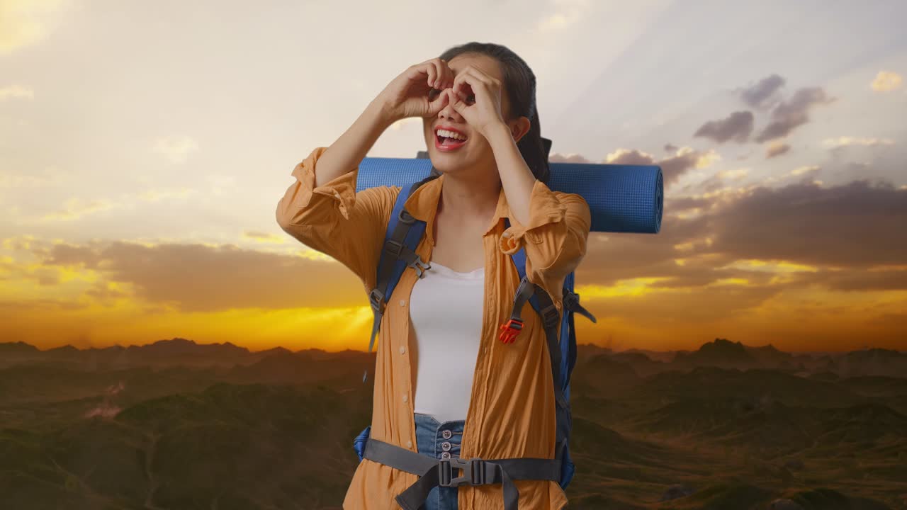 excursionista asiática con mochila de montañismo sonriendo y haciendo gestos de binoculares luego mirando a su alrededor mientras está de pie en la cima de la montaña durante el atardecer