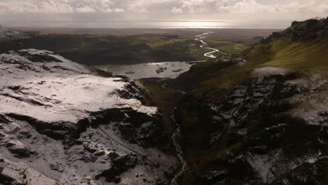 Aerial-view-of-a-snow-dusted-landscape-at-Sólheimajökull,-highlighting-a-winding-river-cutting-through-the-melting-glacier,-with-a-distant-view-of-the-ocean-under-a-cloudy-sky