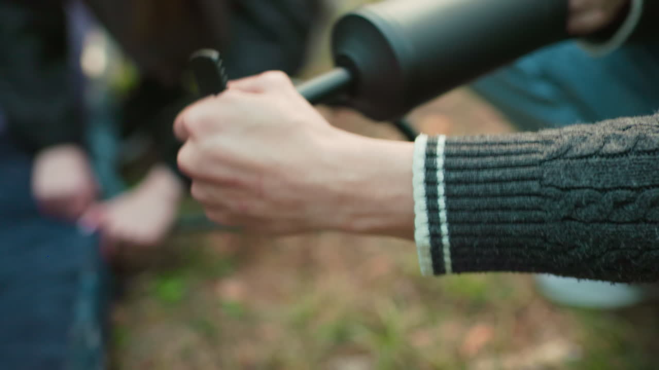 Close up hand view of person using hand pump to inflate object outdoors while another person holds nuzzle in background with blurred forest environment