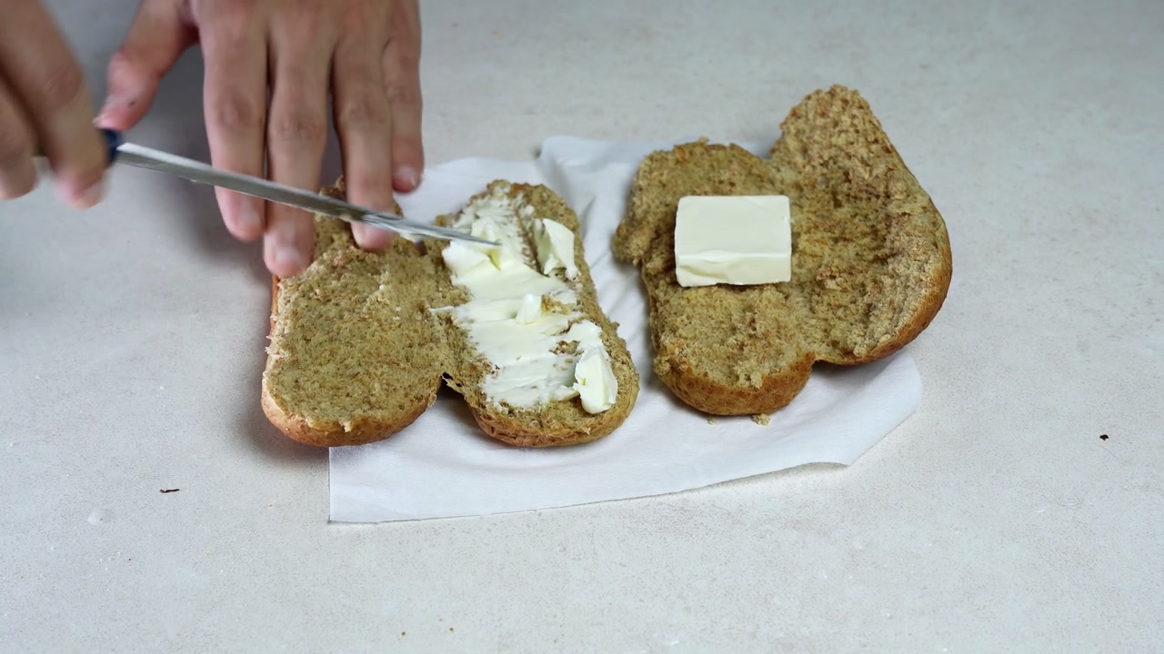 Primo piano dall'alto di mani maschili che spalmano burro morbido su un panino di pane integrale affettato usando un coltello. La preparazione del cibo avviene su un tovagliolo bianco sopra un luminoso bancone da cucina interno.