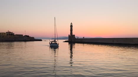 Sail-boat-entering-Chania-Old-Venetian-Port-in-front-of-lighthouse-during-sunset,-Crete