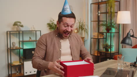 excited young caucasian businessman celebrating birthday opening surprise gift box at table at home