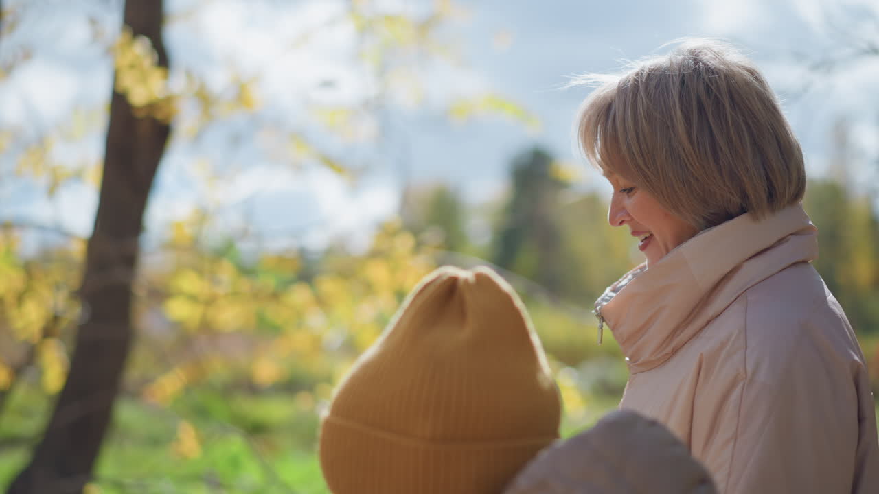 Close up of aunt smiling while walking beside kid in hood and beanie on sunny autumn day, interacting warmly as golden foliage blurs in background with soft sunlight filtering through tree branches