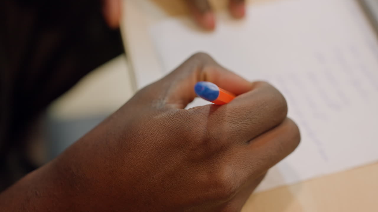 Sorte hænder skriver eksamensnoter på papir med orangeblå kuglepen, anspændt studiestemning, nærbillede af håndskrift og margener, studerende forbereder sig under tidspres i et biblioteks- eller klasseværelsesmiljø.