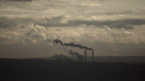 Smoke-billows-from-factory-in-distance-on-gray-cloudy-day,-time-lapse