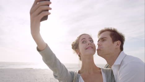 Una-Pareja-Feliz-Tomando-Selfies-Usando-Un-Teléfono-Móvil-En-La-Playa-Al-Atardecer