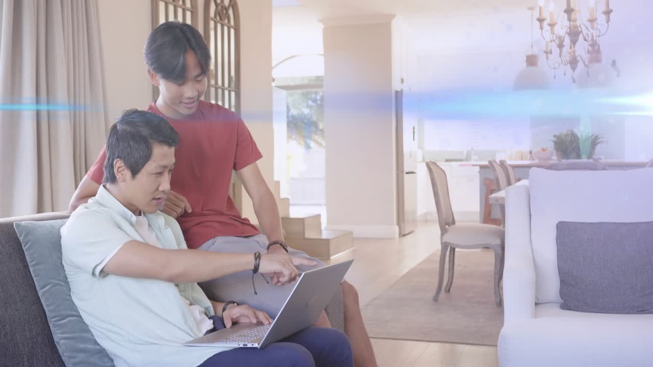 Seated man typing on laptop on couch after teenager leaning in, sharing tech content and smiling