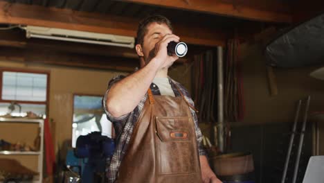 Caucasian-male-knife-maker-in-workshop-standing-at-desk-and-drinking-coffee
