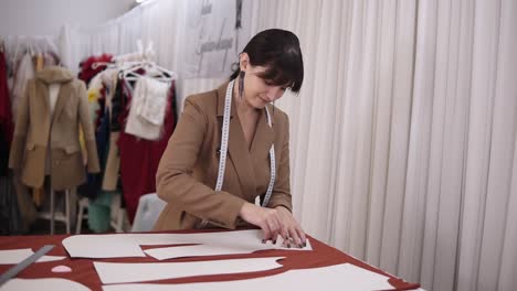 portrait of a young woman in brown suit with a measuring tape on neck drawing the white line on the white cloth, using a white soap or chalk creating pattern of a future dress on the working table in professional studio