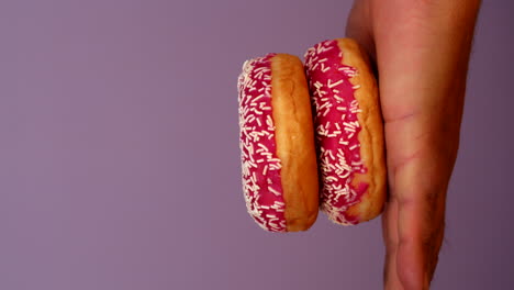 stack of three donuts in male hand. woman putting donut step by step on mans hand. pink doughnut with white sprinkles on pastel light purple background. colorful food.