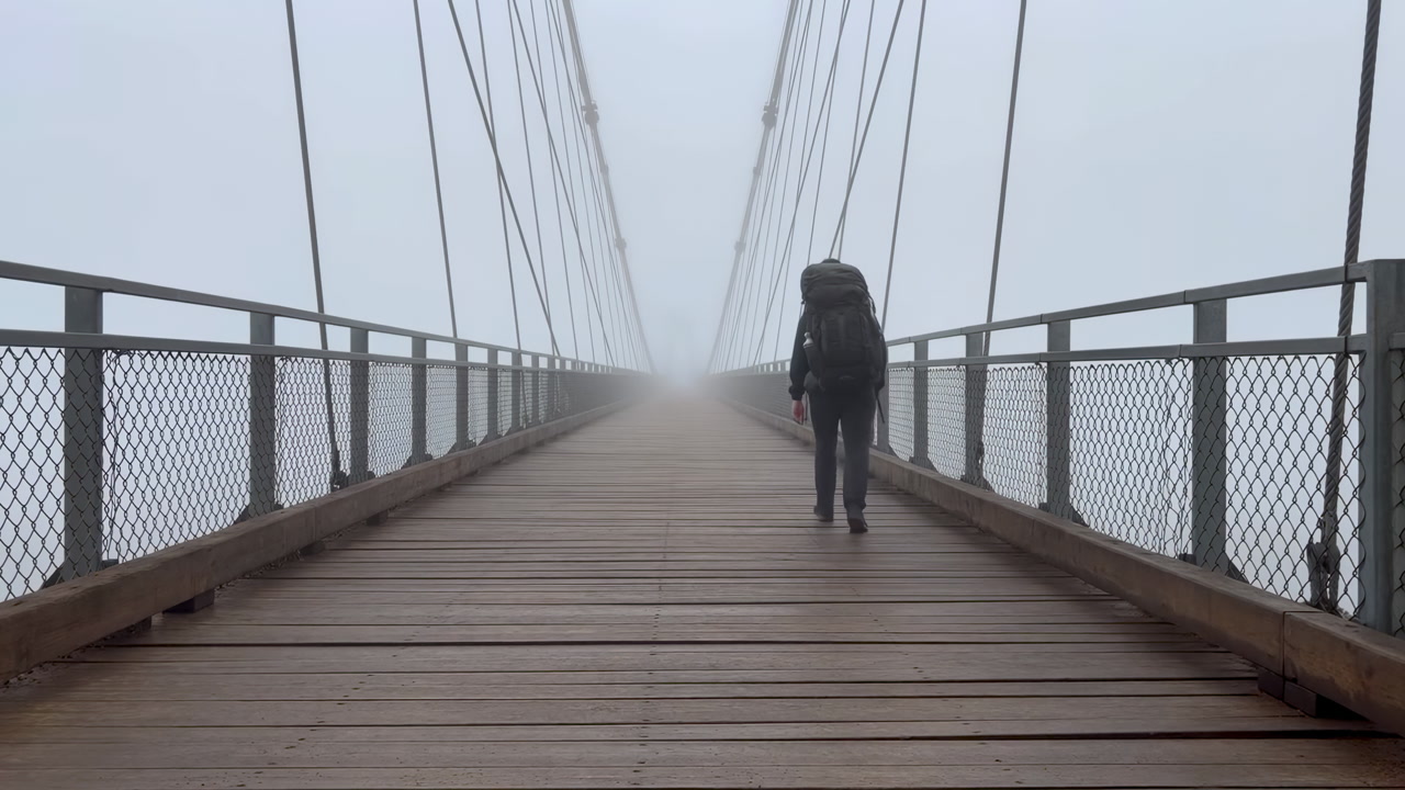 Backpacker Walking Foggy Suspension Bridge Ahead Wooden Planks And Metal Cables Vanish Into Thick Mist
