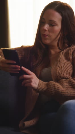 vertical video of woman sitting on sofa at home at streaming or watching movie or show or scrolling internet on mobile phone