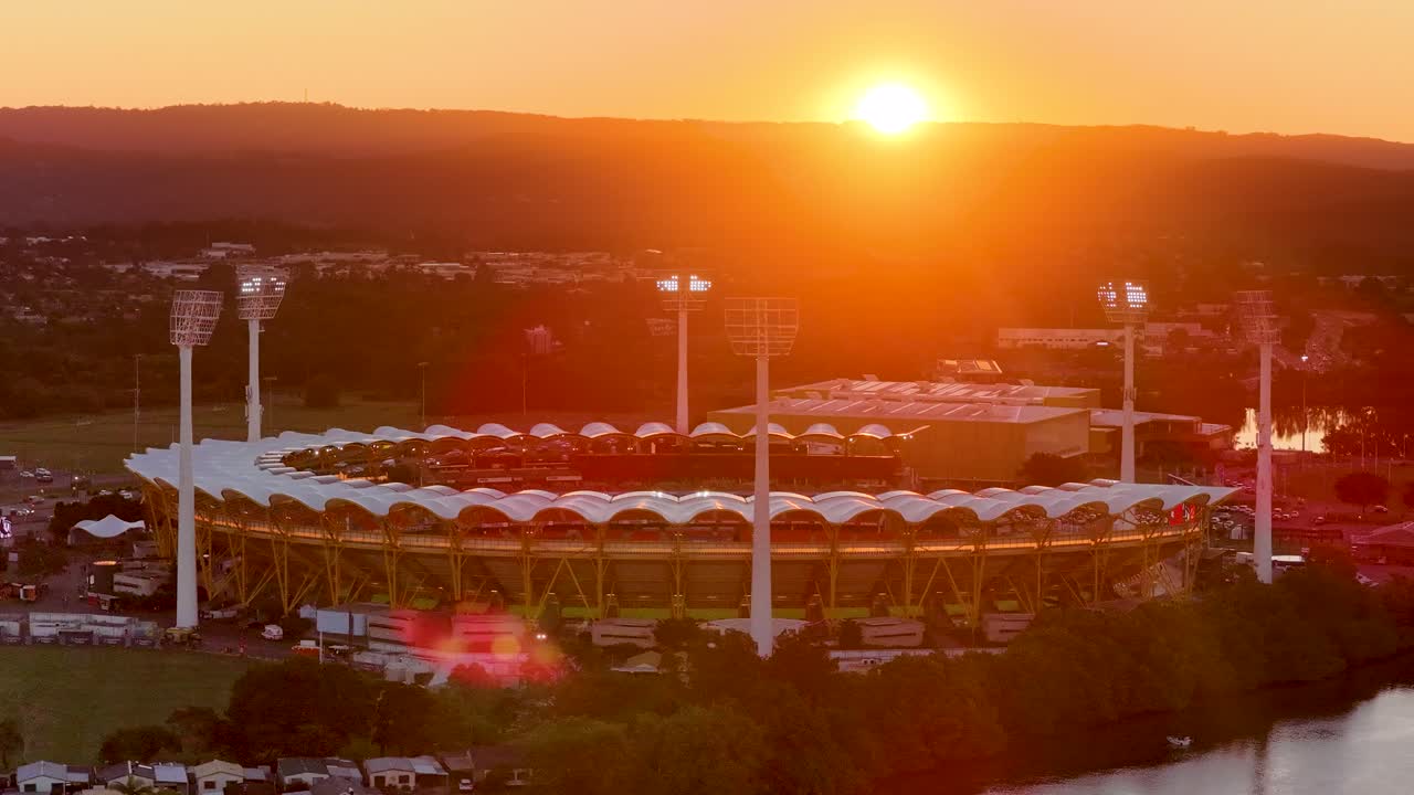 Drone captures a large outdoor stadium at sunset, stadium lights turning on, with river and distant hills in view, creating a dramatic golden atmosphere