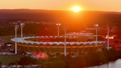 drone captures a large outdoor stadium at sunset, stadium lights turning on, with river and distant hills in view, creating a dramatic golden atmosphere