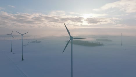 aerial establishing view of wind turbines generating renewable energy in the wind farm, snow filled countryside landscape with fog, sunny winter evening golden hour, wide drone shot moving forward