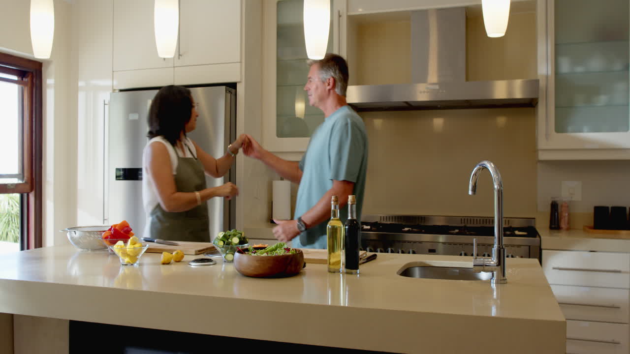 Senior diverse couple dancing joyfully in modern kitchen with fresh salad on counter, at home