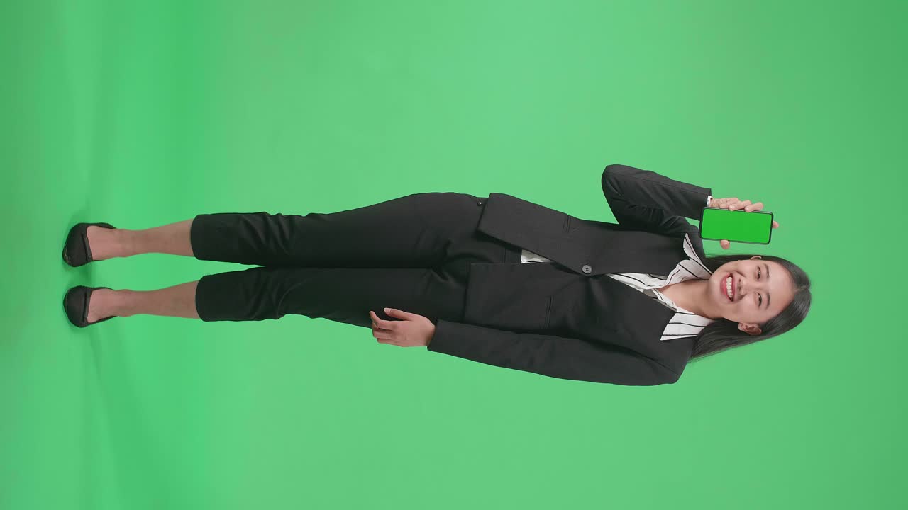 Full Body Of A Smiling Asian Business Woman Holding Green Screen Mobile Phone While Standing In Front Of Green Screen