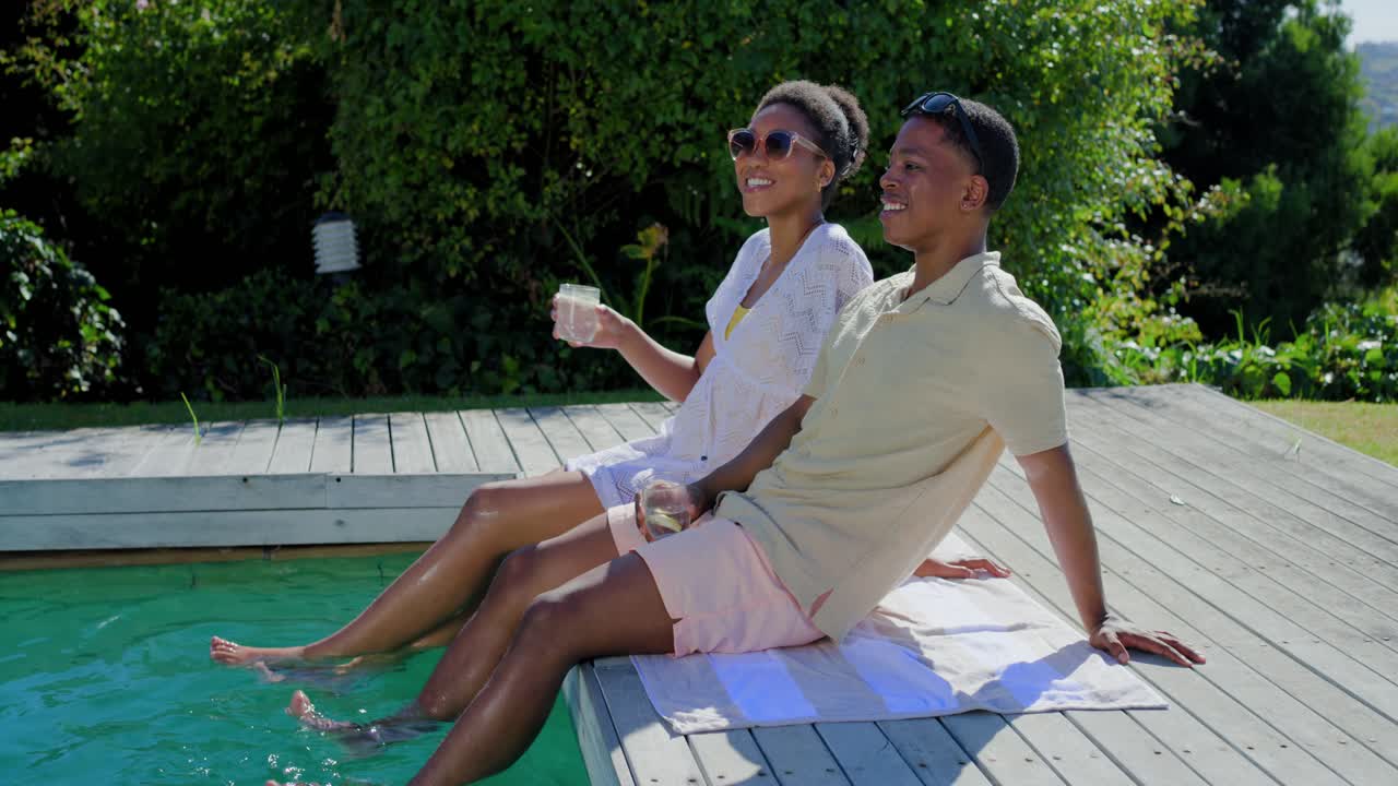 African American couple sitting by pool on deck, dipping feet, holding glasses, in swimwear