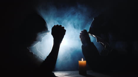 two people sit at a table praying a candle burns near them behind the mist refreshed by a spotlight