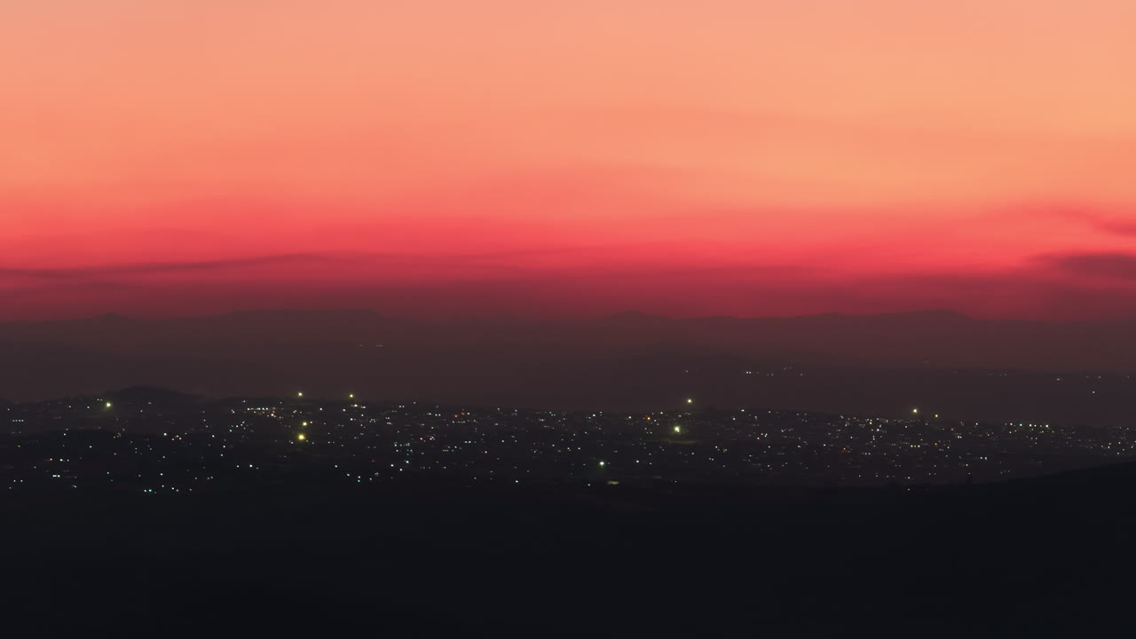 A serene sunset paints the sky in pink and orange hues over distant hills as the lights of nearby towns begin to sparkle