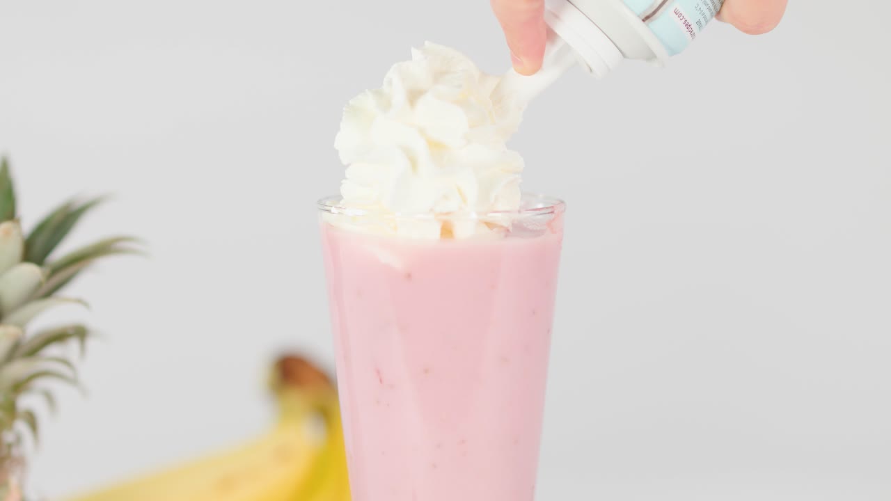 Hand tops creamy pink smoothie with whipped cream, surrounded by fresh fruit, bright studio lighting