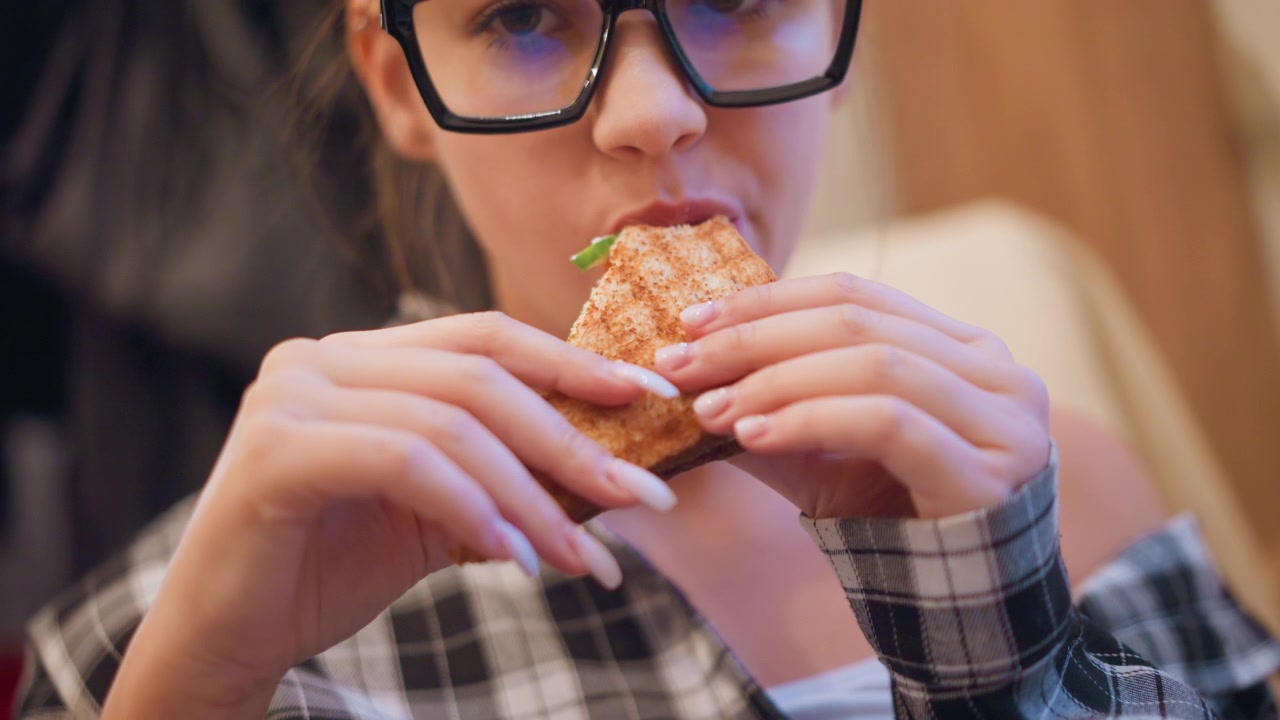 Close up of young woman with long hair and glasses eating food. Woman wears black framed eyeglasses has manicured nails painted white on natural color and is dressed in plaid clothing