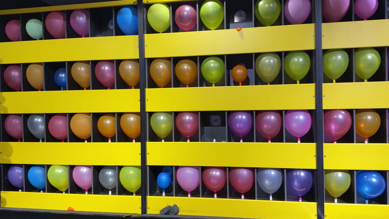 A vibrant array of colorful balloons arranged in rows on a bright yellow carnival balloon popper game wall. Shot indoors under artificial lighting, creating a fun, static recreational background.